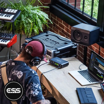 Person working on electronic music equipment in a home studio with a laptop, speakers, and synthesisers, all connected to a MOTU M2 audio interface.